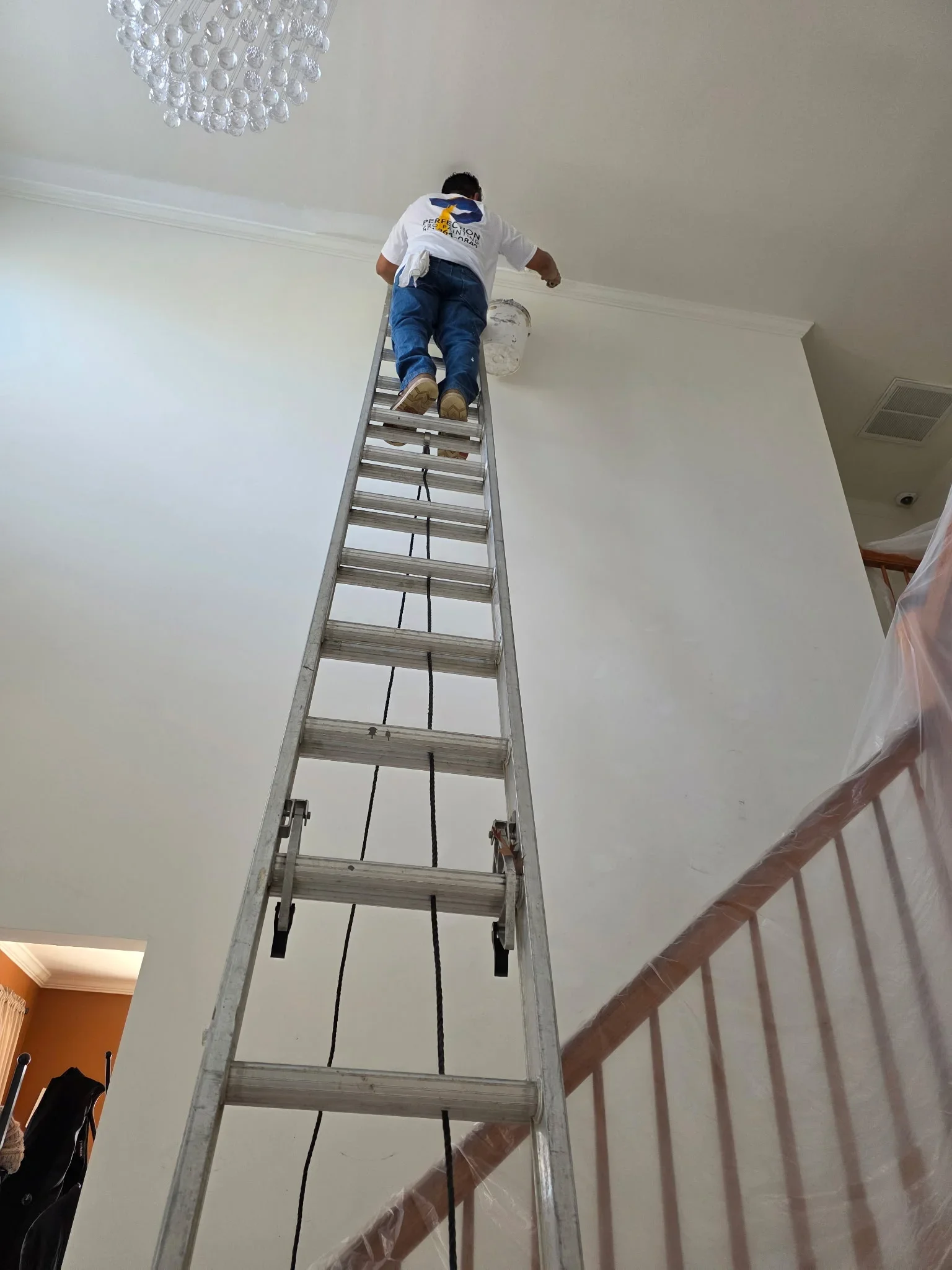 Painter on ladder applying paint to high interior ceiling and walls in residential space