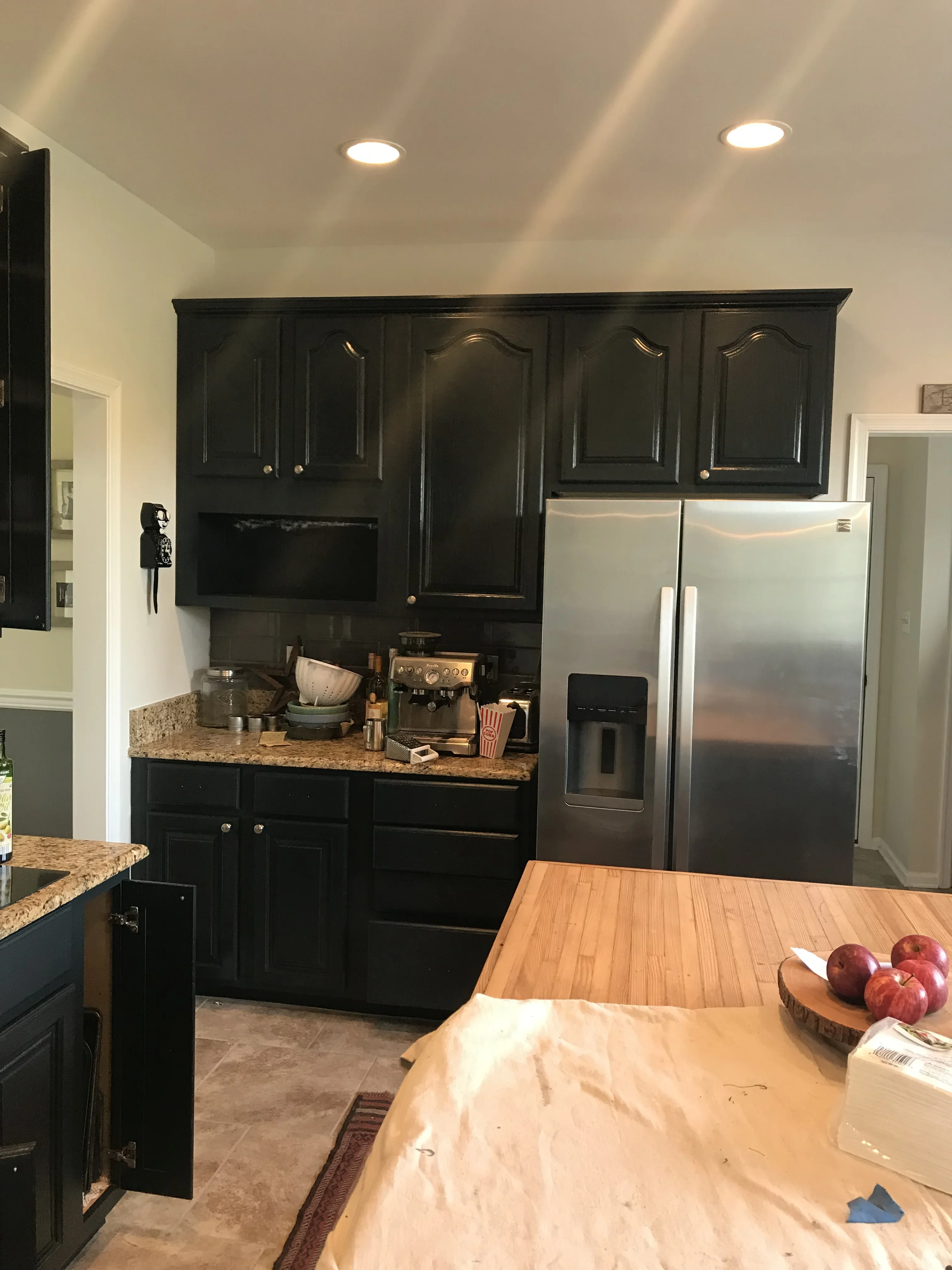 Black painted kitchen cabinets with granite counters, recessed ceiling lights, and stainless steel refrigerator