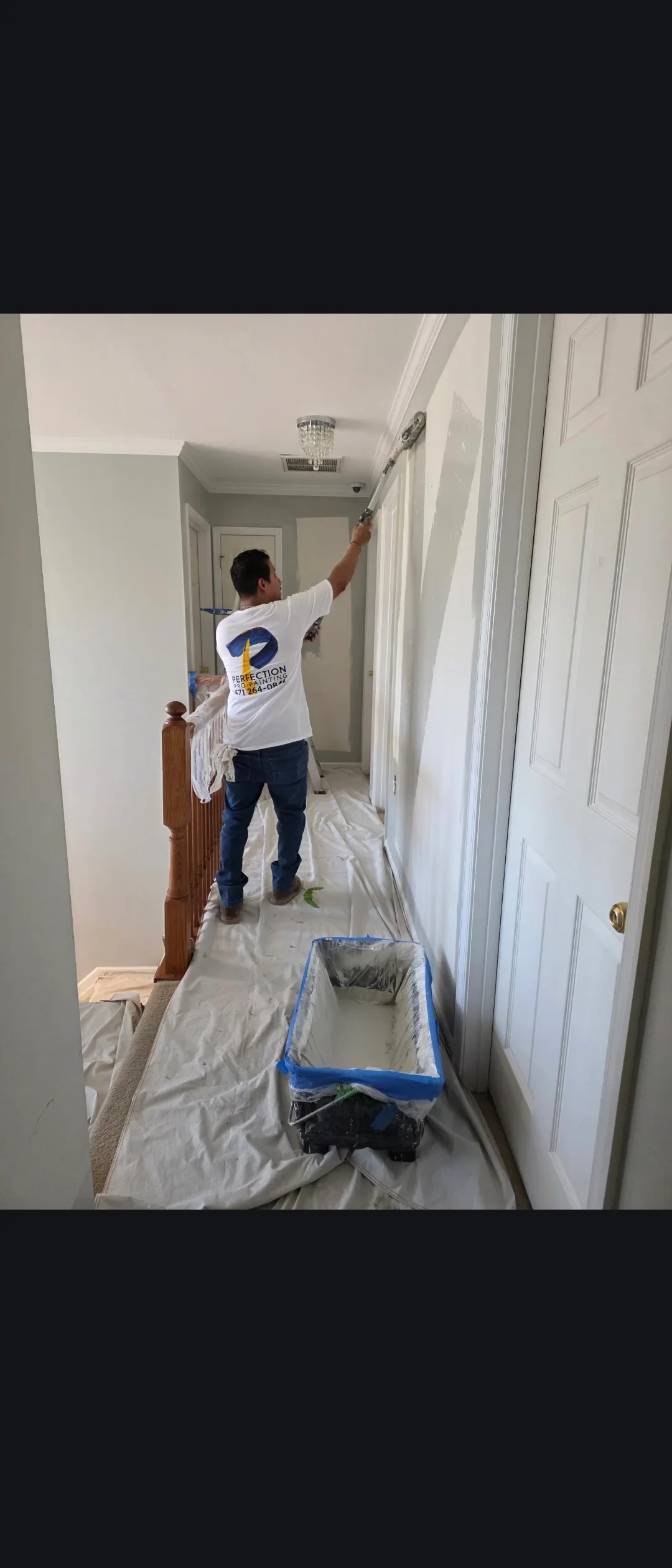 Painter applying white paint to hallway ceiling with white walls and six-panel doors